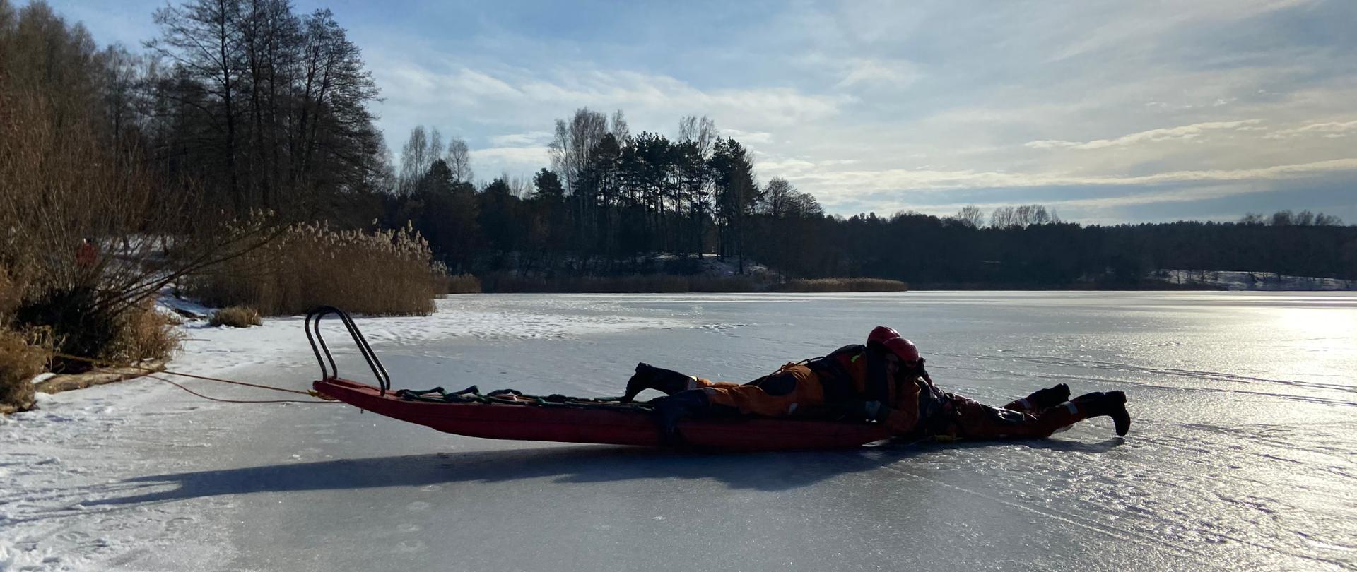 Zdjęcie przedstawia zamarznięty zbiornik wodny a na jego powierzchni (lodzie) znajduje się dwóch strażaków. Jeden na saniach lodowych wyciąga drugiego - symulując załamanie się lodu