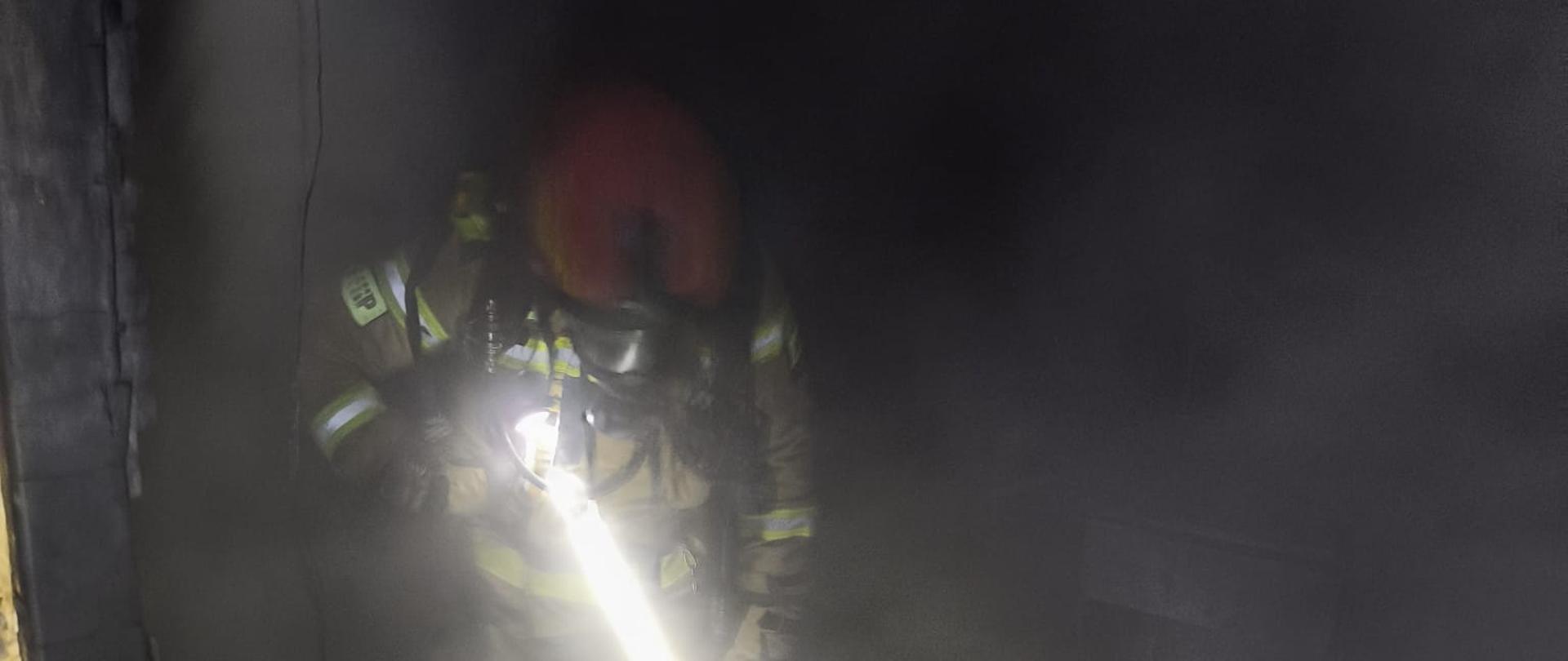 Na zdjęciu widoczny strażak w zadymieniu. Na piersi latarka, widoczny strumień światła. 
