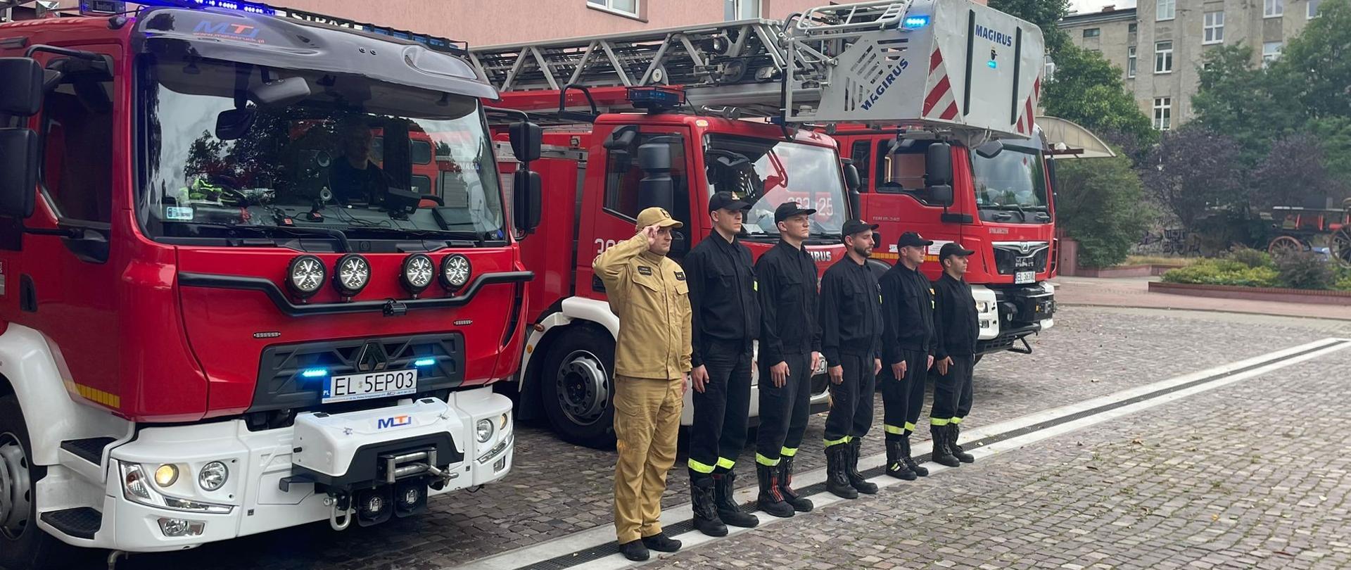 Strażacy ustawieni w rzędzie przed samochodami pożarniczymi oddają hołd poległym w powstaniu warszawskim