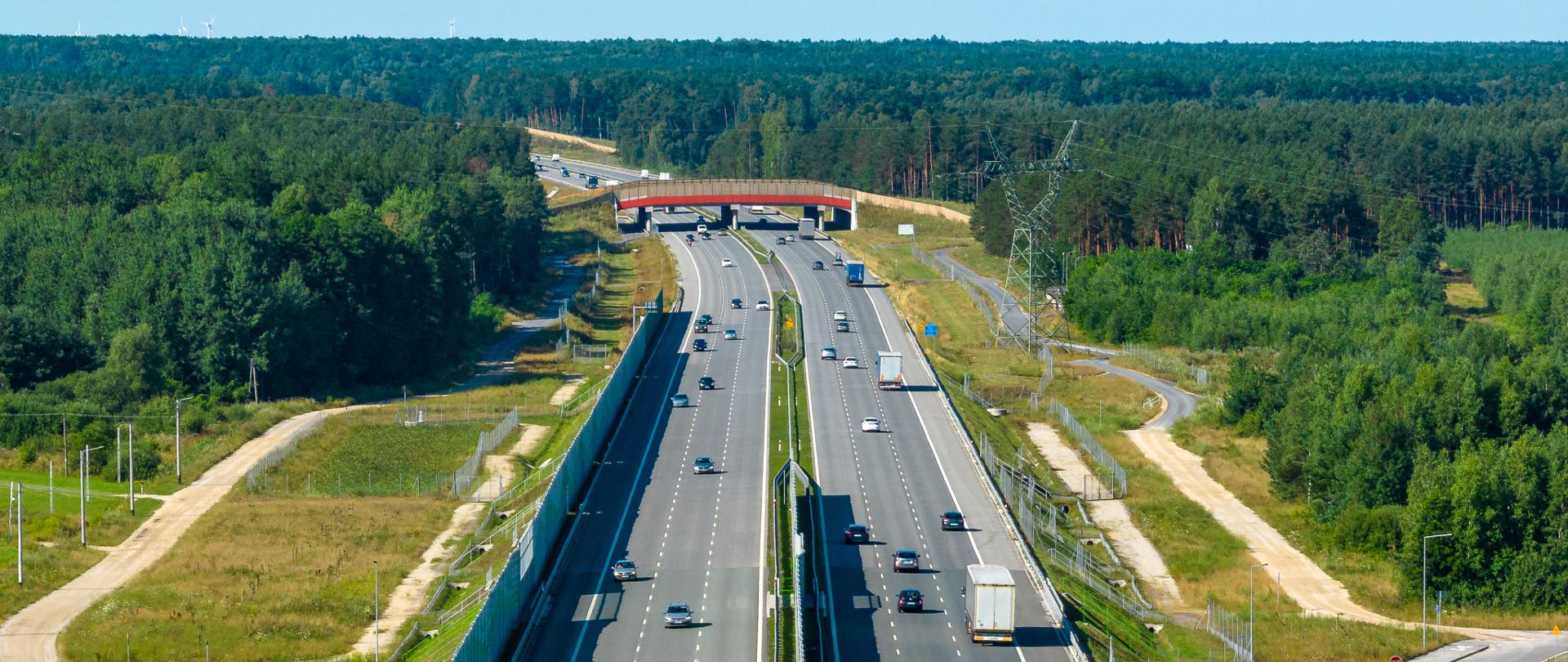 Ruch pojazdów na autostradzie A1 w okolicach Radomska. Trzy pasy ruchu w każdą stronę. Środkowej części zdjęcia nad autostradą znajduje się przejście dla zwierząt. Po obu stronach drogi rośnie gęsty las.