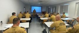 Odprawa szkoleniowa dla koordynatorów prewencji społecznej z KP/M PSP woj. opolskiego. Strażacy Państwowej Straży Pożarnej w umundurowaniu służbowym siedzą na sali szkoleniowej i słuchają wykładu prelegenta.