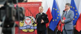 Mężczyzna w strażackim mundurze generalskim oraz drugi w okularach i szarym garniturze stojący przed mikrofonami. Za nim flagi Polski oraz Unii Europejskiej oraz logo Państwowej Straży Pożarnej. Na pierwszym planie z lewej strony widoczna kamera.
