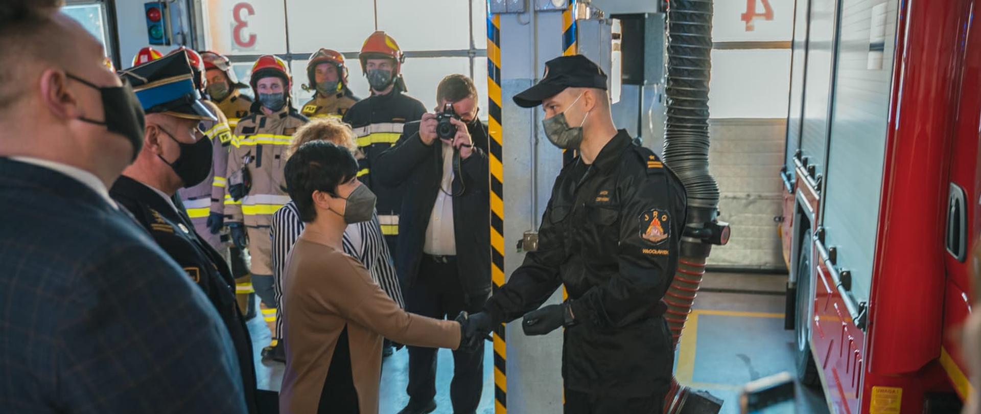 Pani Poseł gratuluje dowódcy nowego auta. W tle załoga i goście. Rzecz dzieje się w garażu.