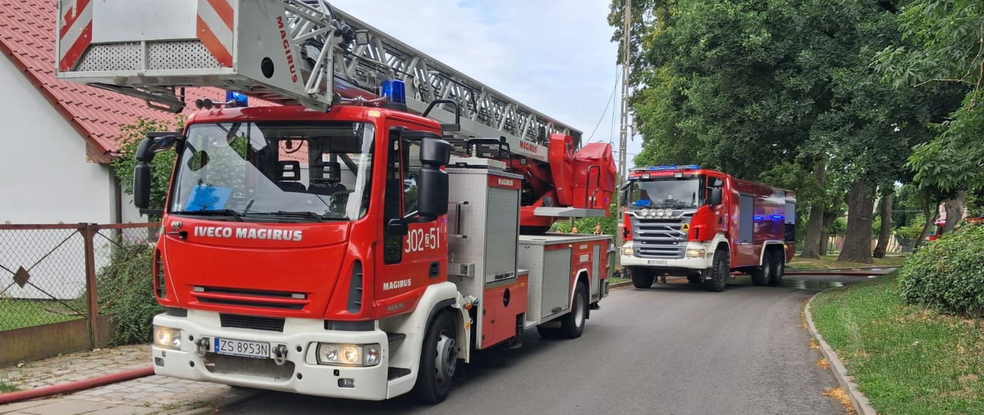 Na asfalcie, na lekkim łuku drogi, widać dwa czerwone samochody strażackie jadące jeden za drugim. Pierwszy, bliżej obiektywu, to pojazd z drabiną mechaniczną marki Iveco Magirus. Ma czerwoną kabinę z białym zderzakiem, a na górze złożoną konstrukcję metalowej drabiny ze znakiem "MAGIRUS". Na drzwiach kabiny widać złote numery taktyczne „302[Z]51” oraz napis „MAGIRUS”. Tablica rejestracyjna ma numer „ZS 8953N”. Za nim, w pewnej odległości, jedzie drugi, większy samochód gaśniczy, również marki Iveco, z widocznymi niebieskimi światłami ostrzegawczymi. Po lewej stronie drogi stoi dom z czerwonym dachem i siatkowym ogrodzeniem, po prawej widać szpaler dużych, liściastych drzew i trawnik.