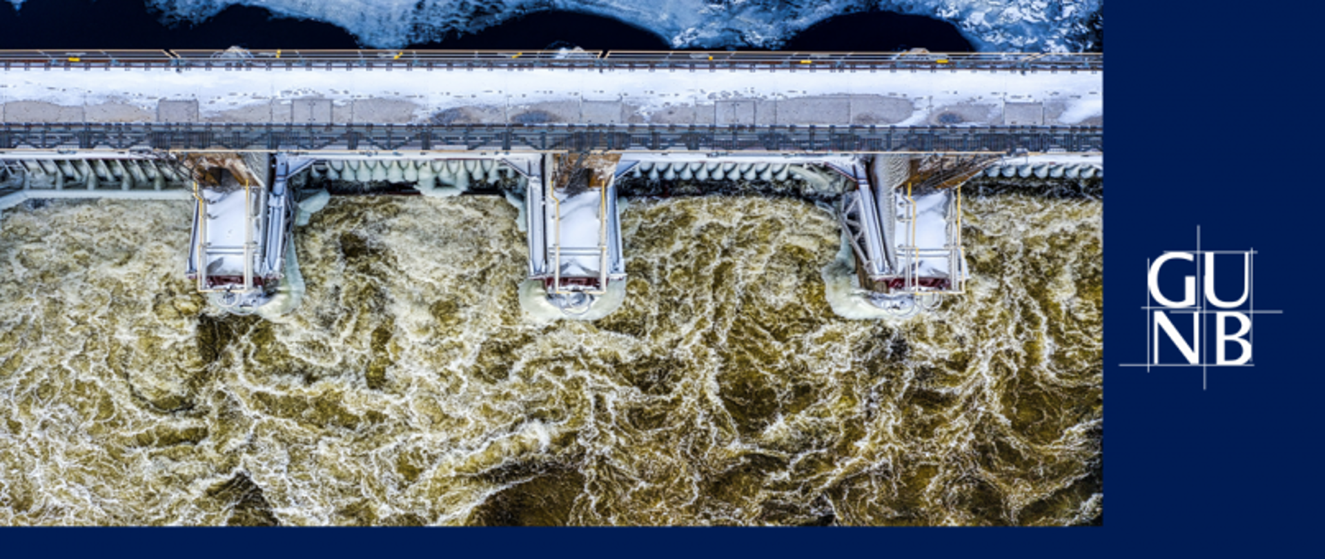 Zapora wodna zimą z otwartymi śluzami i spływającą wodą; widok z góry, śnieg na konstrukcji.