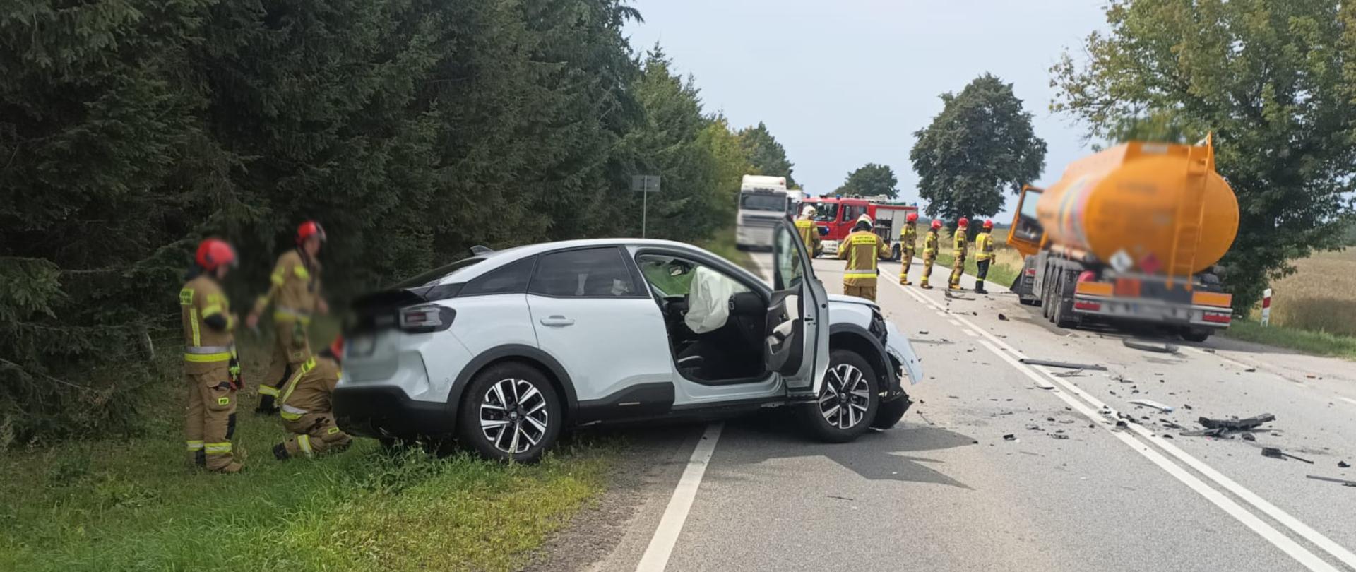 Na zdjęciu wykonanym na jezdni drogi krajowej DK10 na pierwszym planie znajduje się uszkodzony po wypadku pojazd osobowy. Przy pojeździe strażacy PSP w trakcie czynności. Na drugim planie na skraju jezdni cysterna przy, której strażacy również prowadzą czynności służbowe.