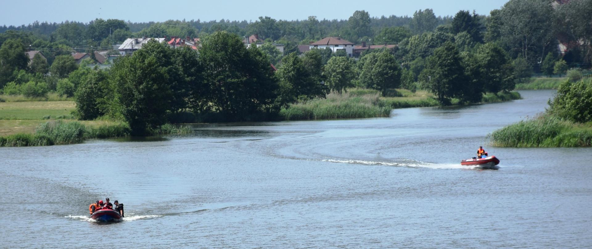 Dwie łodzie płynące po zbiorniku wodnym. W tle, na brzegu rosnące drzewa i stojące budynki.