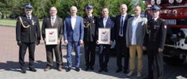 osiem osób pozuje do zdjęcia pamiątkowego, trzy osoby ubranych w mundury galowe a pięć u ubraniach cywilnych, dwie trzymają przed sobą pamiątkowe dyplomy w tle rosną liczne drzewa, po prawej stronie na pierwszym planie pojazd ciężarowy straży pożarnej