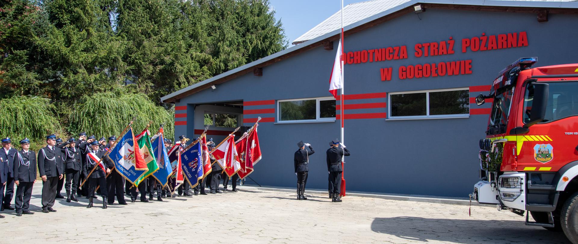Po lewej stronie stoi szpaler strażaków, część z nich trzyma sztandary, w centralnej części stoi trzech strażaków stanowiących poczet flagowy i następuje wciągnięcie flagi, po prawej stronie stoi samochód ratowniczo-gaśniczy Tło zdjęcia tworzy budynek remizy z napisem "OHOTNICZA STRAŻ POŻARNA W GOGOŁOWIE.