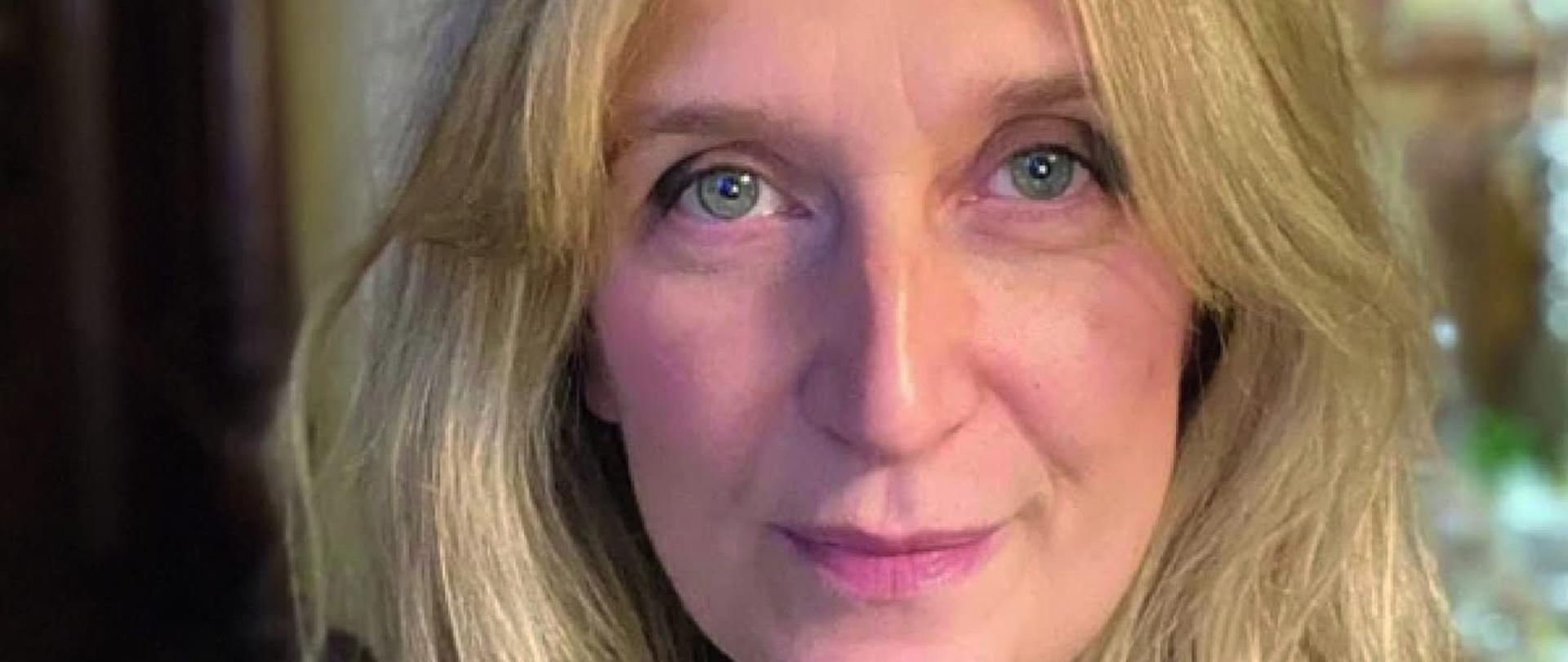 Portret kobiety w średnim wieku. Kobieta ma blond włosy do ramion. Przedstawiona od piersi w górę, w ciemnym swetrze. Tło rozmyte