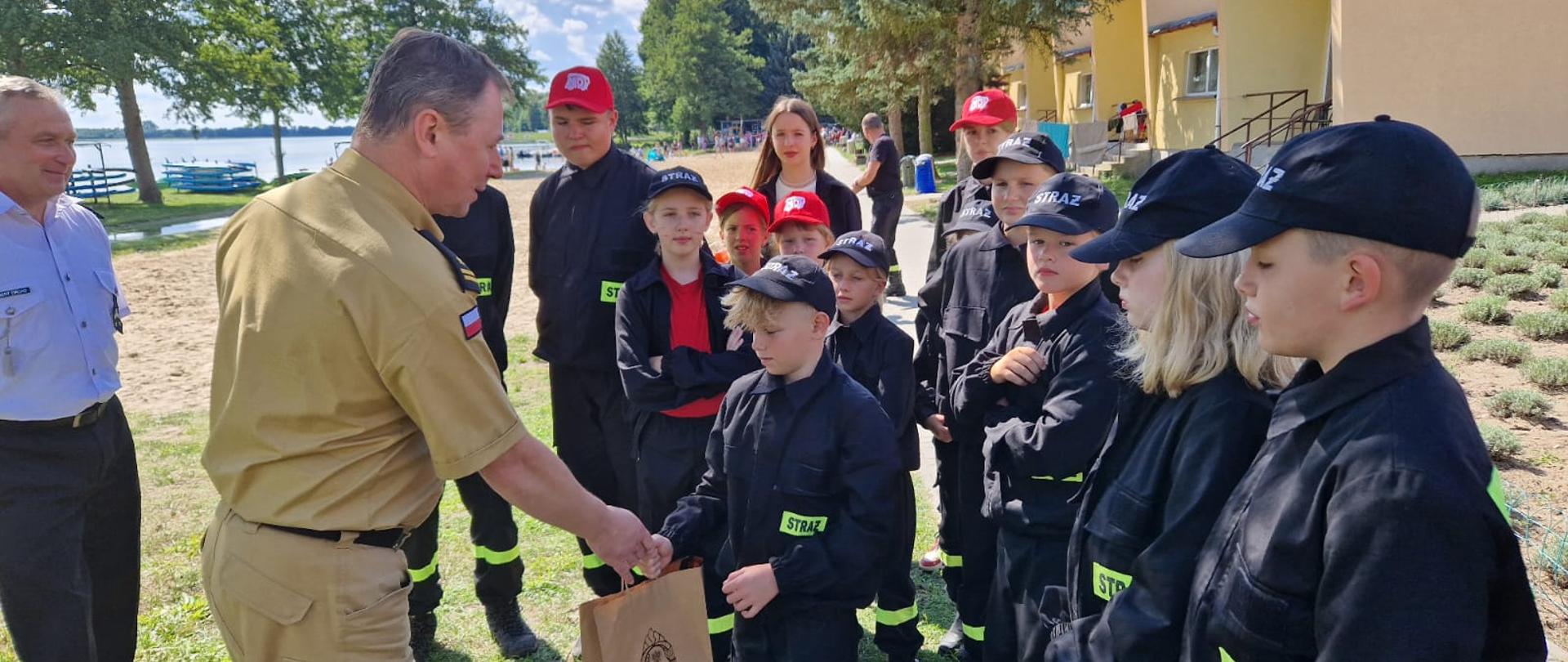 Zdjęcie przedstawia Komendanta Powiatowego PSP w Wolsztynie podczas wizyty na obozach MDP w Wieleniu. Na zdjęciu widać grupę dzieci w mundurkach strażackich oraz przedstawicieli PSP wręczających podarunki dla uczestników obozu. Zdjęcie wykonane na zewnątrz, ładna pogoda.