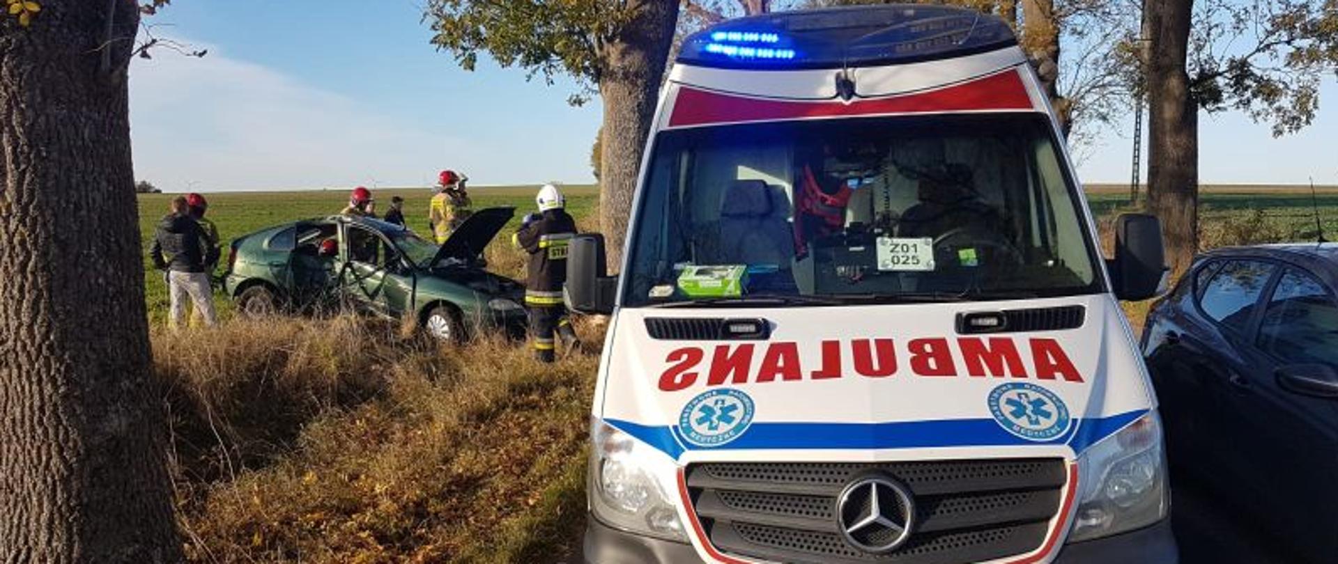 Centralny punkt zdjęcia stanowi karetka pogotowia . W tle po lewej stronie rozbite auto. Przy aucie stoją strażacy.