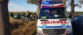 Centralny punkt zdjęcia stanowi karetka pogotowia . W tle po lewej stronie rozbite auto. Przy aucie stoją strażacy.