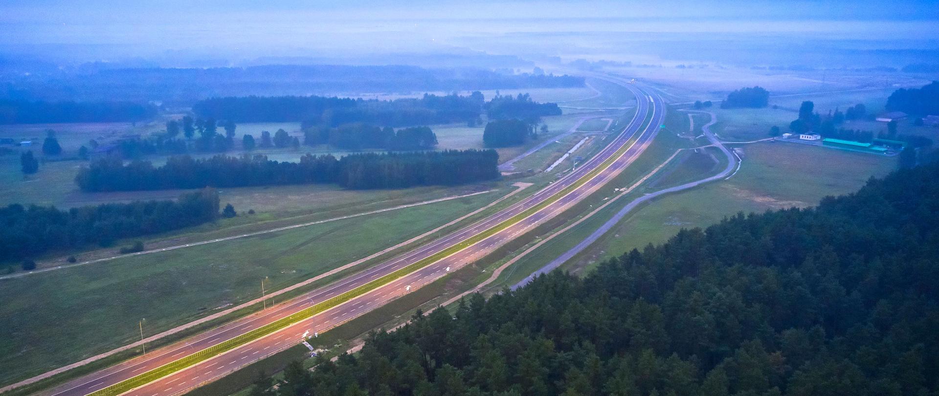 Widok z góry na drogę ekspresową. Wschód słońca, widać unoszące się mgły. Po obu stronach drogi lasy. Na drodze widać jadące auta.