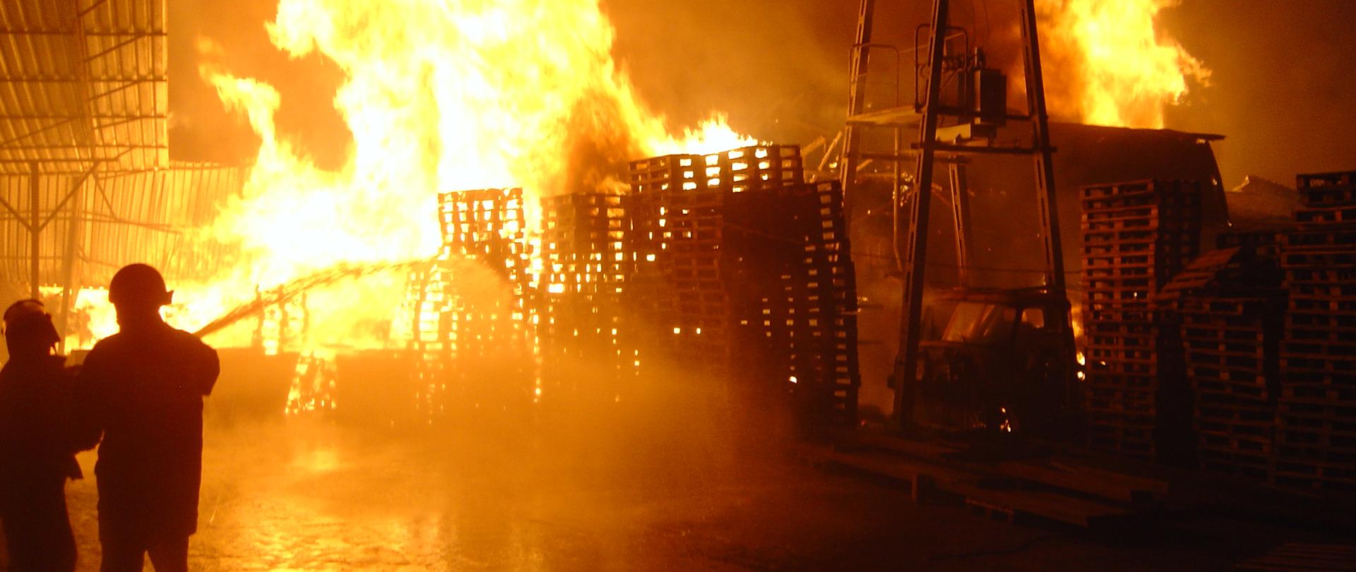 Na zdjęciu widać pożar palet ułożonych w stosy. Po lewej stronie widoczny jest fragment wiaty, pod którą ułożone zostały palety. Dwóch strażaków podaje prąd wody na ogień sięgający kilku metrów. 