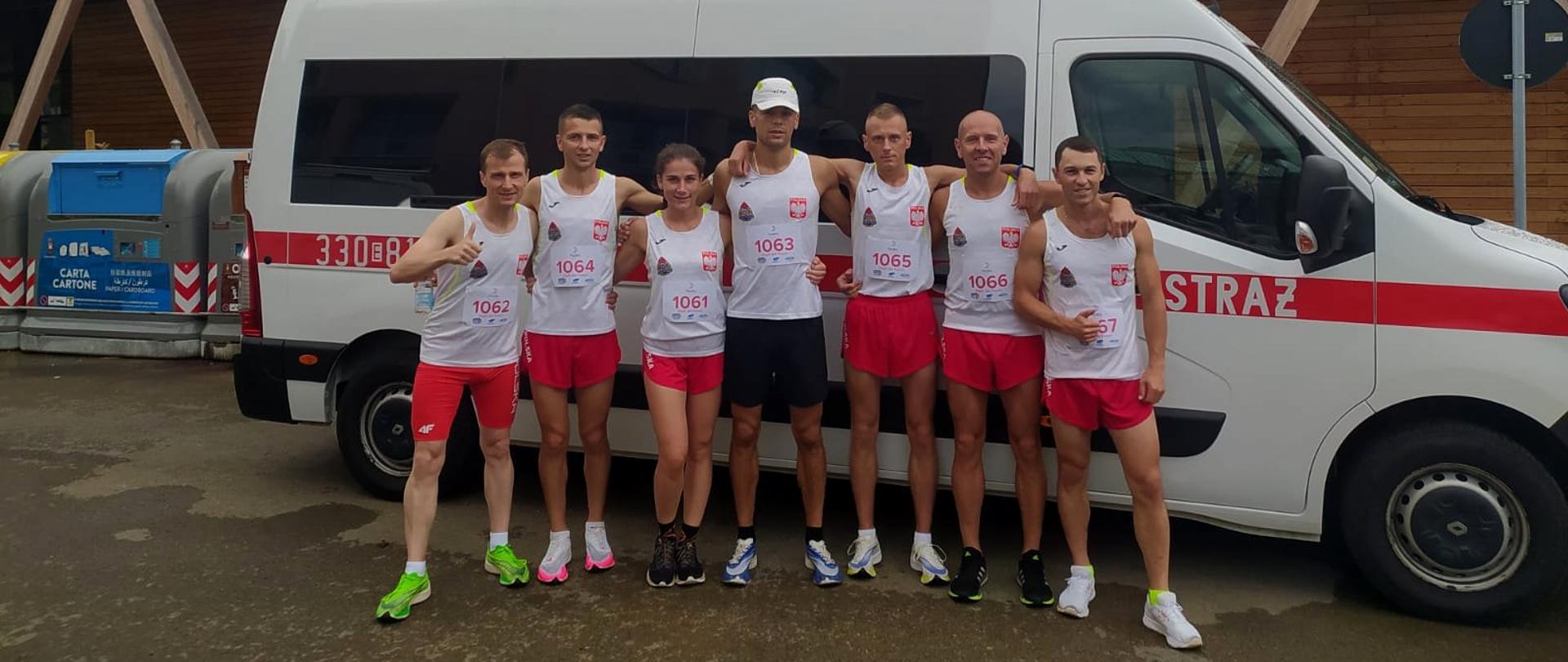 Zdjęcie przedstawia 7 osób biegaczy- 6 mężczyzn i 1 kobieta stojących na tle oznakowanego jako straż białego samochodu. Wszyscy biegacze ubrani w białe koszulki z godłem i logiem PSP. 