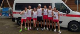 Zdjęcie przedstawia 7 osób biegaczy- 6 mężczyzn i 1 kobieta stojących na tle oznakowanego jako straż białego samochodu. Wszyscy biegacze ubrani w białe koszulki z godłem i logiem PSP. 