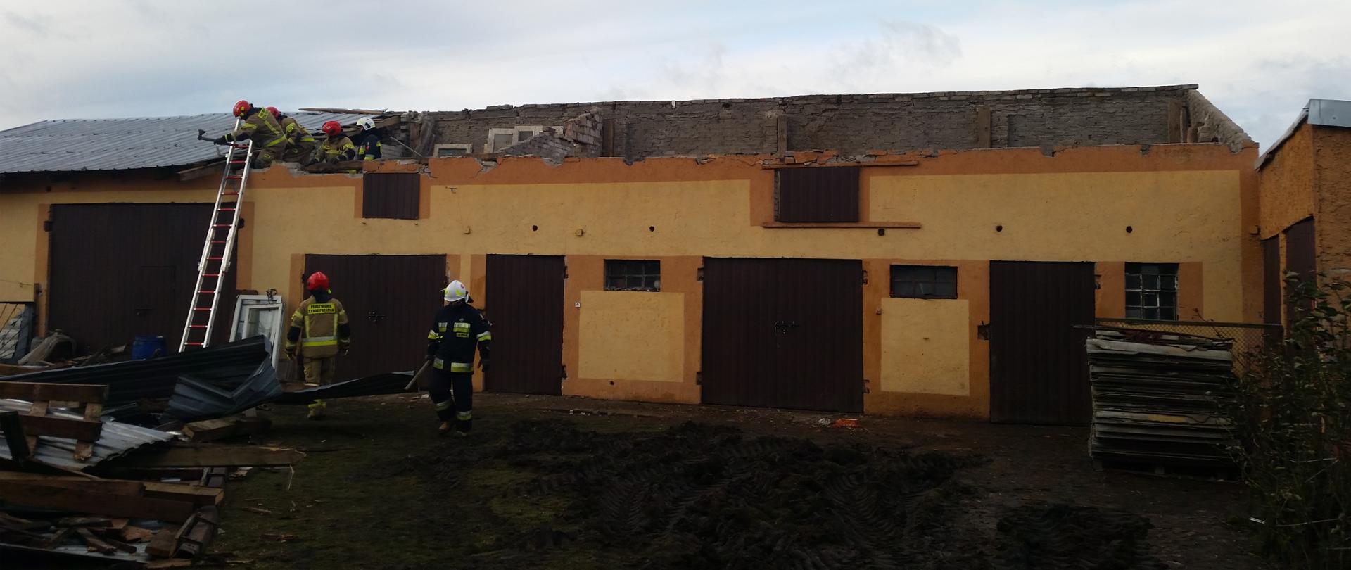 Strażacy PSP i OSP Pracują przy uszkodzonym budynku gospodarczym, z którego wiatr zerwał dach.