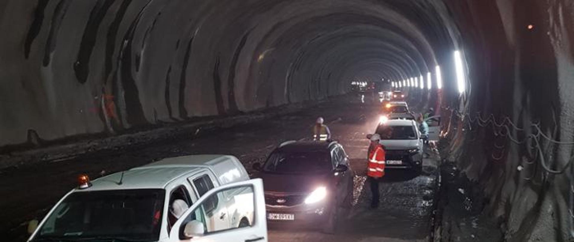 Tunel drogowy, wewnątrz panuje półmrok. Na pierwszym planie widoczne 3 auta, dwie osoby i światła. 