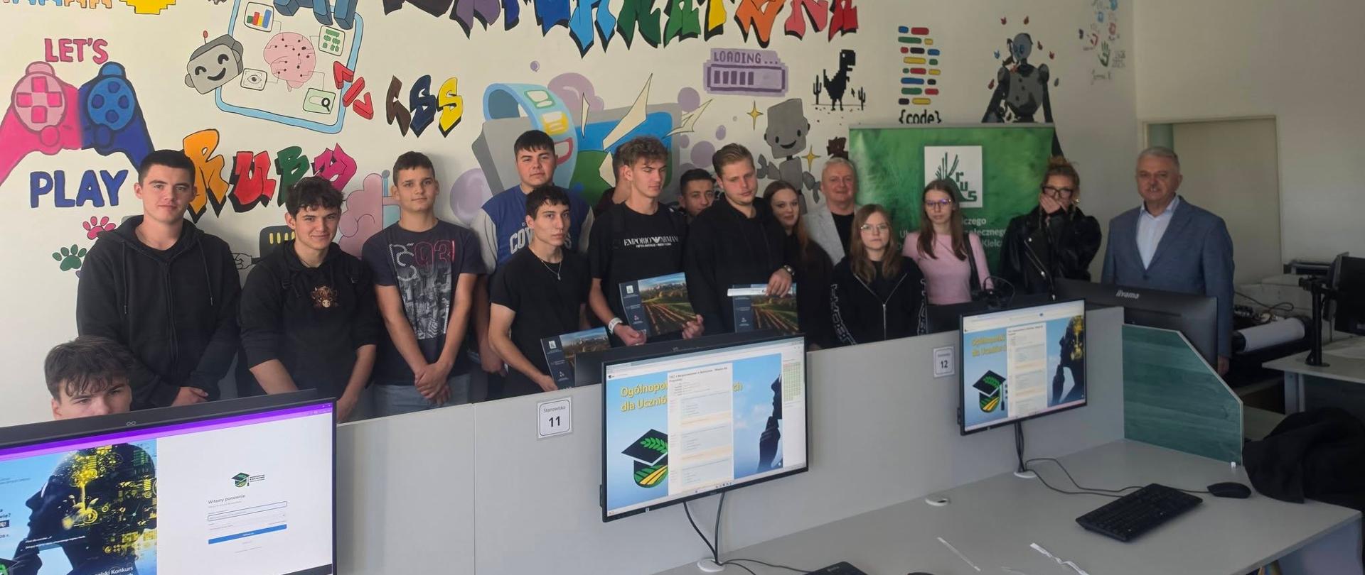 Zdjęcie przedstawia grupę uczestników zgromadzonych w nowoczesnej pracowni komputerowej. W pierwszym planie widoczne są stanowiska komputerowe z monitorami wyświetlającymi stronę dotyczącą ogólnopolskiego konkursu dla uczniów szkół rolniczych. W tle grupa pozuje do wspólnego zdjęcia na tle dużego, kolorowego muralu z napisem „AI Informatyka” oraz grafikami związanymi z technologią, programowaniem, sztuczną inteligencją i grami komputerowymi. Część uczestników trzyma materiały lub nagrody konkursowe. Po prawej stronie znajduje się zielony baner KRUS