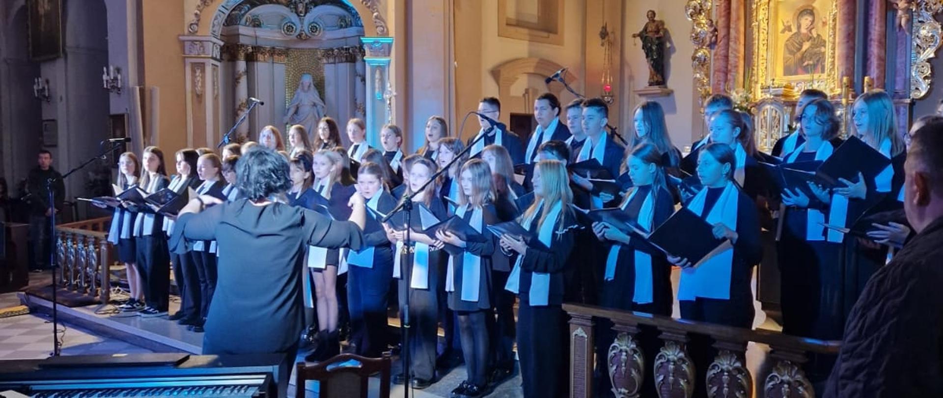 Zdjęcie przedstawia występ chóru we wnętrzu kościoła. Uczniowie ubrani są w jednolite stroje (czarne ubrania z białymi szalami). Chórem dyryguje osoba stojąca z przodu, tyłem do wiernych. W lewym dolnym rogu widoczny jest instrument klawiszowy i krzesło. W prawym dolnym rogu widoczna jest mała dziewczynka z dużą białą kokardą na głowie, obserwująca występ. Całość rozświetlona jest kolorowymi światłami scenicznymi.