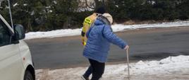 Zdjęcie przedstawia starszą kobietę z laska oraz pomagającego jej strażaka, który trzyma ja pod pachę. Idą po zaśnieżonym chodniku. W tle widzimy drogę, dom, drzewa. W lewym kadrze widać białego busa. 