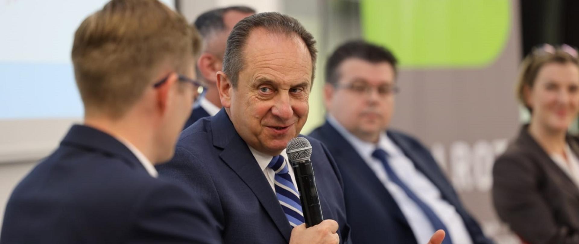 Wiceminister rozwoju i technologii Andrzej Gut-Mostowy podczas obrad konferencji z okazji Światowego Dnia Turystyki odbywającej się w Chełmie, wiceminister siedzi i trzyma w ręku mikrofon, obok uczestnicy spotkania