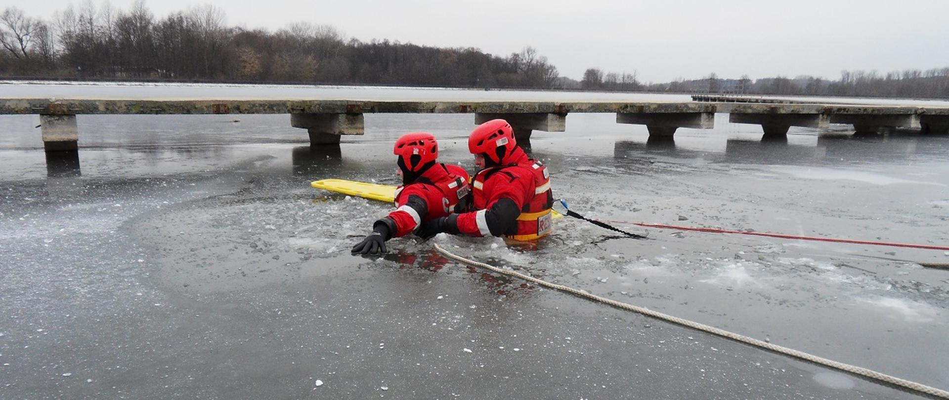 Zdjęcie przedstawia dwóch strażaków ćwiczących na zamarzniętym zbiorniku wodnym z wykorzystaniem deski ortopedycznej