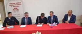 Zdjęcie przedstawia moment podpisywania umowy na dofinansowanie sprzętu przy stole prezydialnym przez Prezesa OSP Gnojnica Wola oraz Zastępcy Prezesa WFOŚiGW w Rzeszowie Bogusława Kidę. Na zdjęciu za stołem prezydialnym siedzą od lewej: Komendant Powiatowy PSP w Ropczycach, Poseł na sejm Kazimierz Moskal, Prezes OSP Gnojnica Wola dh Kacper Darłąk, Zastępca Prezesa Zarządu WFOŚiGW w Rzeszowie Bogusław Kida oraz Starosta Powiatu ropczycko-sędziszowskiego Witold Darłak.