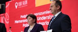 Wiceminister rozwoju i technologii Andrzej Gut-Mostowy i minister odpowiedzialna za turystykę Bułgarii Stela Baltova trzymają w dłoniach dokument z podpisanym porozumieniem o współpracy między MRiT a Ministerstwem Turystyki Republiki Bułgarii, za ich plecami czerwony baner z logo TOUR SALON

