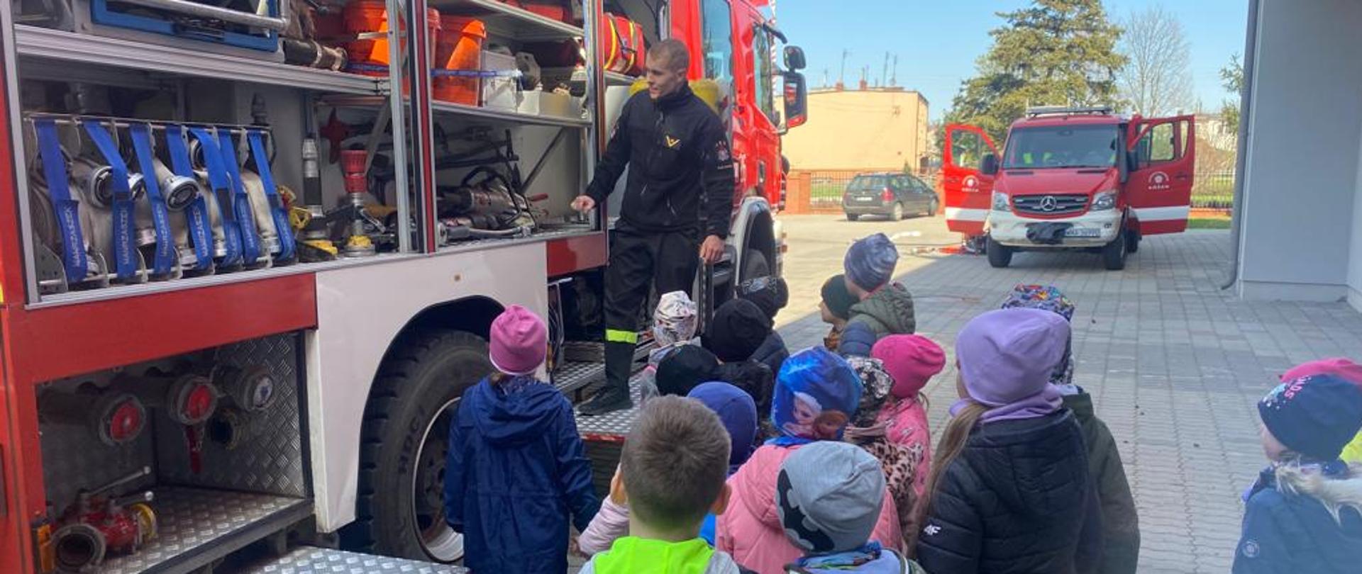 Strażak stoi przed samochodem strażackim i pokazuje sprzęt będący na wyposażeniu dzieciom stojącym przed pojazdem.