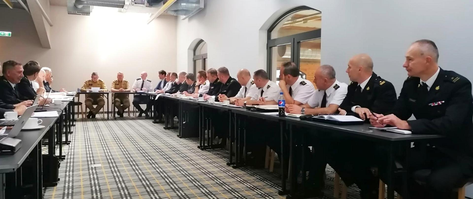 na zdjęciu dwudziestu strażaków siedzących w czasie narady przy stołach ustawionych w kształt litery U, dwunastu ubranych jest w mundur wyjściowy, sześciu w białe koszule z krótkimi rękawami, dwóch w nowe umundurowanie koloru piaskowego, na podłodze wykładzina z wyraźnymi drobnymi pasami na suficie instalacja wentylacyjna