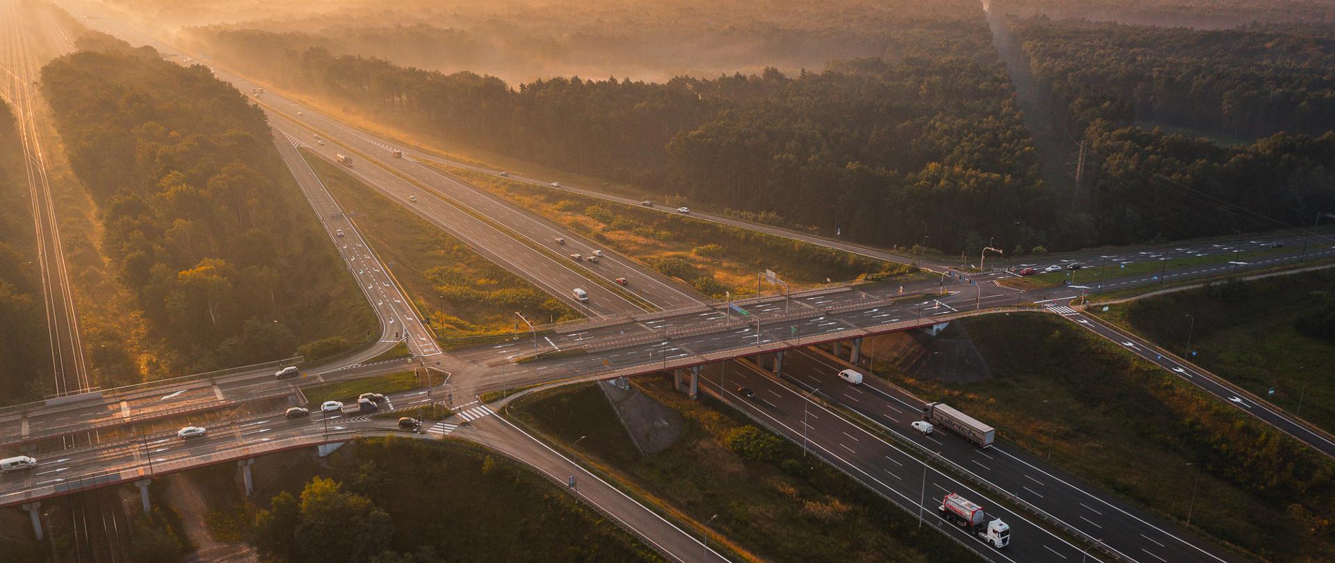 Widok z lotu ptaka na węzeł drogowy Zabrze Południe na autostradzie A4 o wschodzie słońca. Przez środek kadru przebiega wielopasmowa jezdnia z samochodami osobowymi i ciężarowymi. Nad autostradą poprowadzono drogę poprzeczną na wiadukcie. Wokół znajdują się rozległe tereny leśne, a w tle widoczna jest poranna mgła i zabudowa Górnośląsko-Zagłębiowskiej Metropolii.