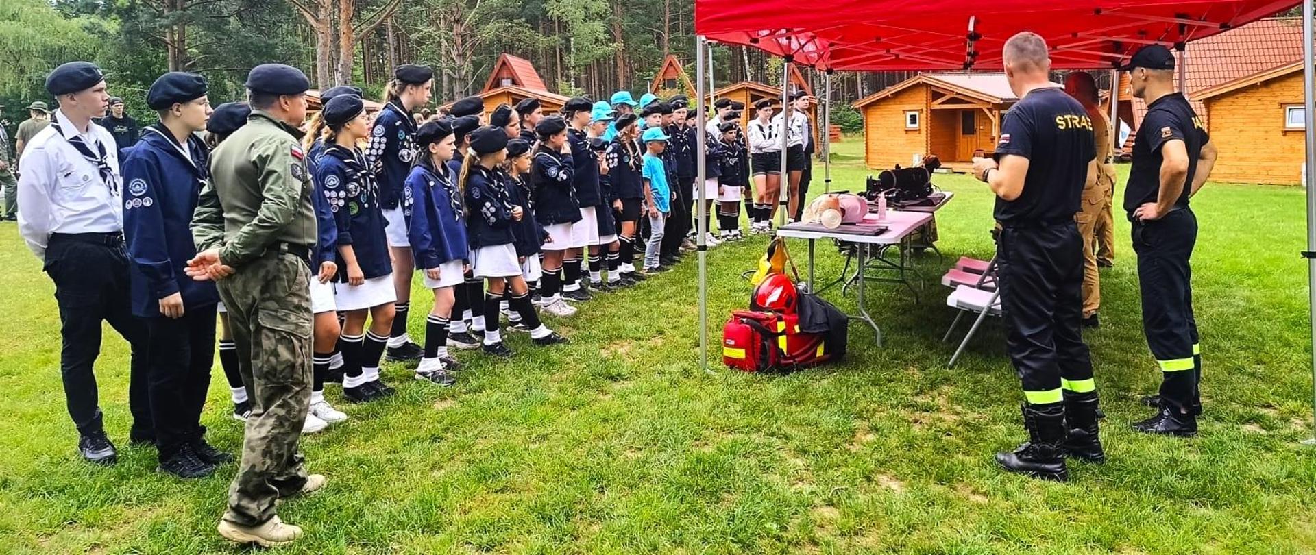 Grupa harcerzy stoi w rzędzie przy czerwonym namiocie. Dzieci są ubrane w mundury harcerskie. Część dzieci ma ubranie koloru granatowo-białego oraz granatowe czapki. Część dzieci ma ubrane błękitne koszulki, błękitne czapki i ciemnoniebieskie spodnie. Dziewczynki mają ubrane białe spódnice i długie skarpetki. Pod namiotem stoją strażacy. Dwóch strażaków jest ubranych w mundury koloru czarnego. Obok widać dwóch strażaków ubranych w umundurowanie koloru piaskowego. Strażacy stoją przy stoliku. Na stoliku widać sprzęt strażacki. Przy stoliku widać na ziemi czerwoną torbę i czerwony kask. W tle widać drewniane domki i las. 