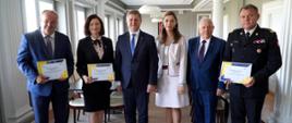 Zdjęcie zrobione na sali konferencyjnej. Sześć osób stoi w szeregu, trzy z nich trzyma pamiątkowe dyplomy. Po prawej stronie stoi oficer Państwowej Straży Pożarnej w stopniu generała.