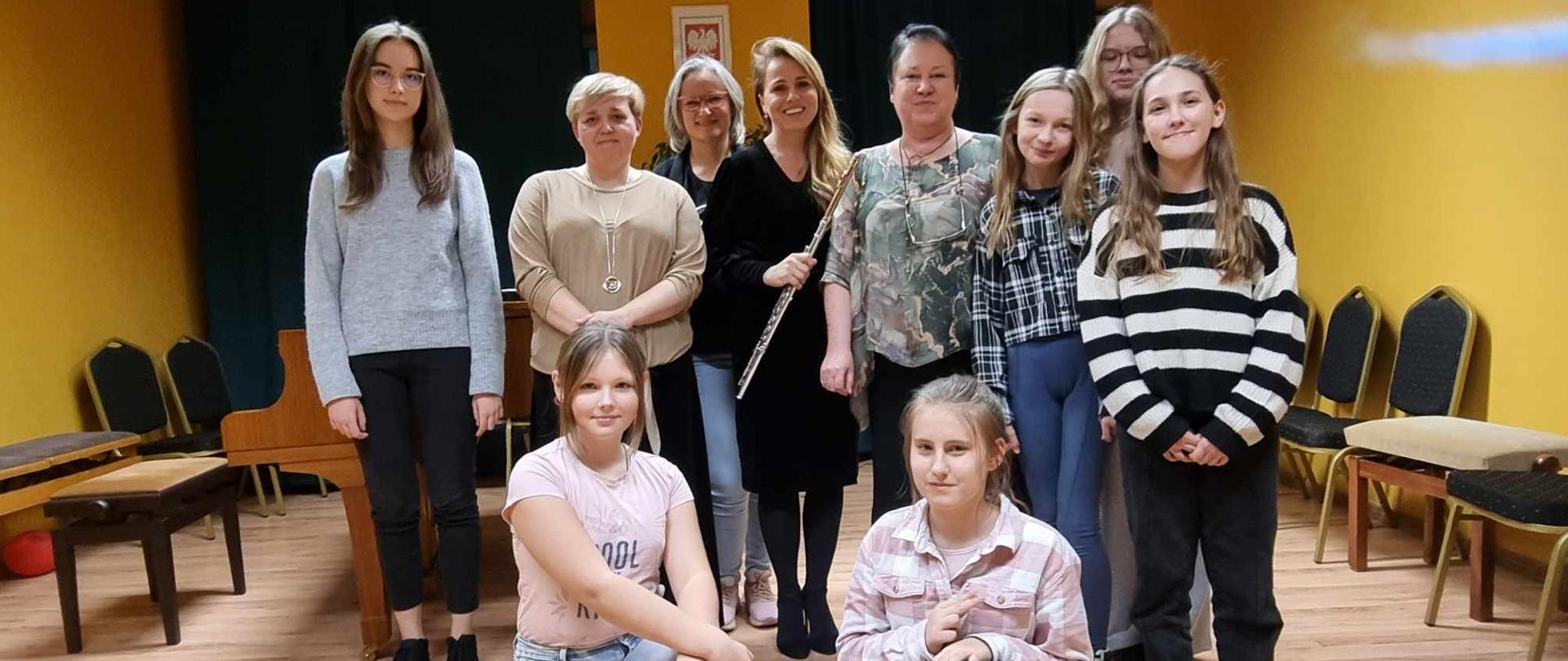 Zdjęcie grupowe. dwie dziewczyny kucają. 8 kobiet stoi za nimi. Kobieta stojąca po środku w czarnej sukience trzyma w dłoniach flet poprzeczny