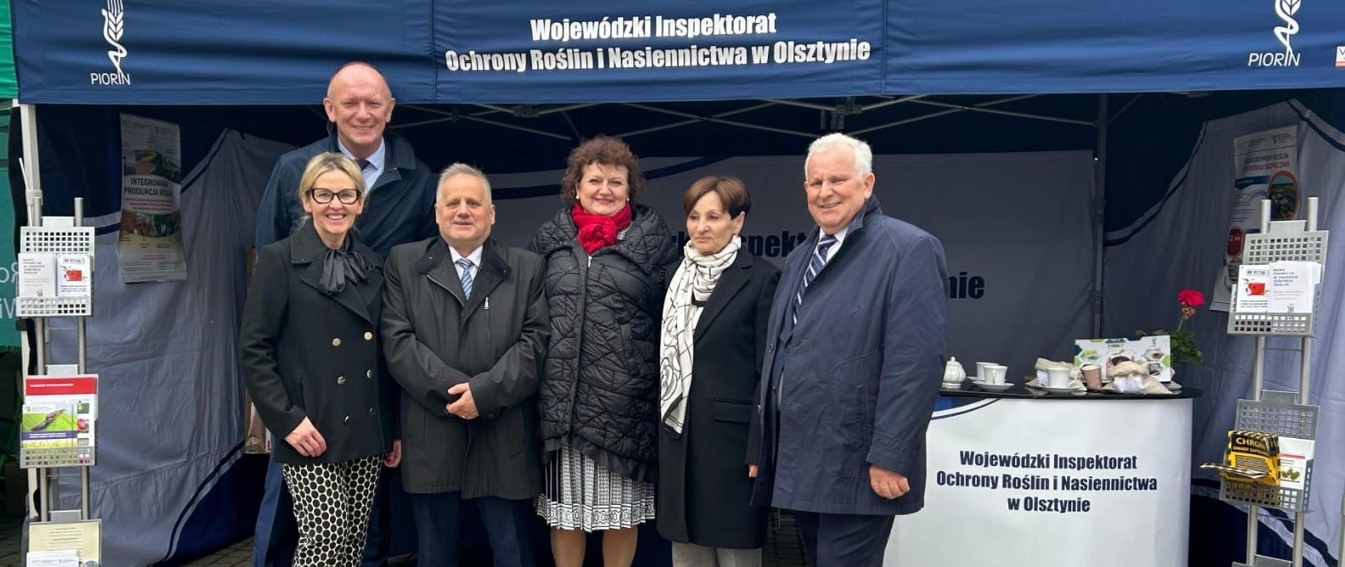 Grupa osób pozuje do zdjęcia na tle stoiska WIORiN, Wojewódzka Inspektor Katarzyna Dębowska, za nią WIJHARS Ryszard Piórkowski, Wicewojewoda Warmińsko-Mazurski Zbigniew Szczypiński, WIIH Krystyna Procyk, Wojewódzka Inspektor Maria Paszko i Jan Paszko
