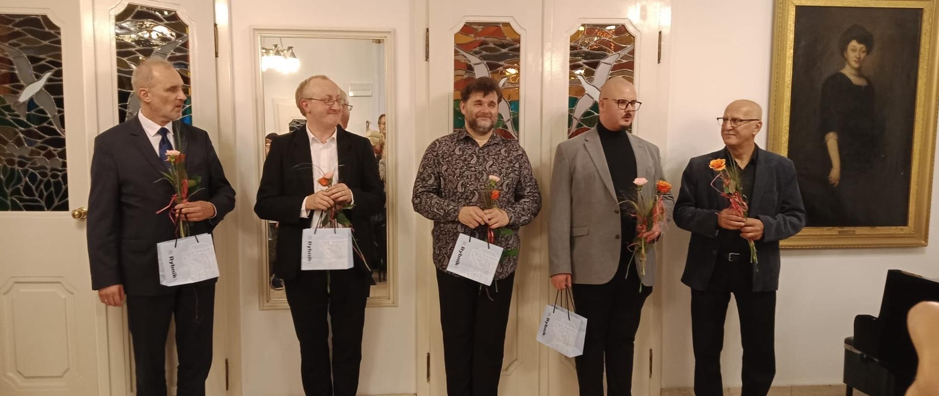 Kolorowe zdjęcie, na którym w jednym rzędzie stoi 5 wykonawców (od lewej: Franciszek Wieczorek, Artur Hes, Adam Mokrus, Adam Dzierżęga, Adam Abrachamczyk). Panowie ubrani w koncertowe stroje, trzymają w rękach kwiatki i rybnickie torebki podarunkowe.