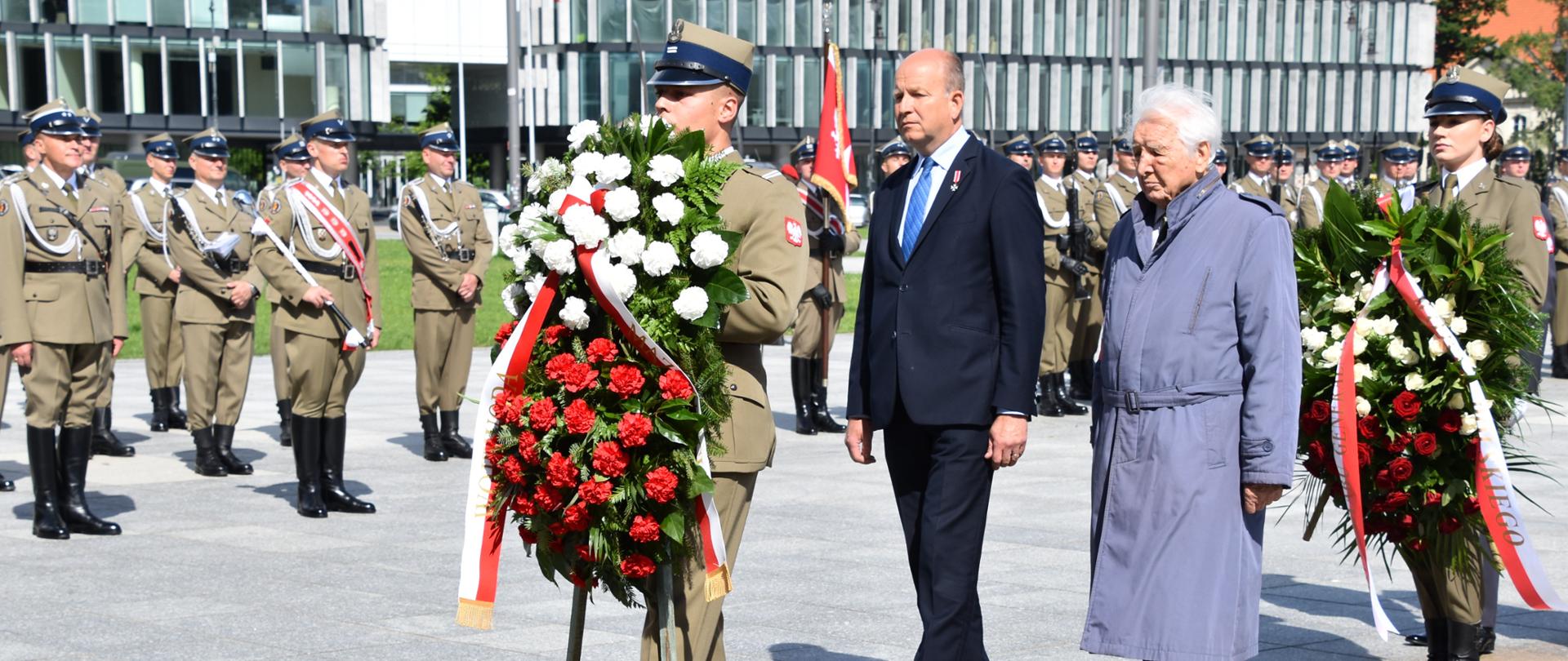 Na pierwszym planie zdjęcia znajduje się czterech mężczyzn, z których dwaj ubrani są w mundury Wojska Polskiego i trzymają w rękach wieńce z biało-czerwonymi goździkami. Pozostali mężczyźni ubrani są - jeden w ciemny garnitur, zaś drugi w długi błękitny płaszcz. W tle znajdują się ustawieni w szpaler żołnierze Wojska Polskiego. 