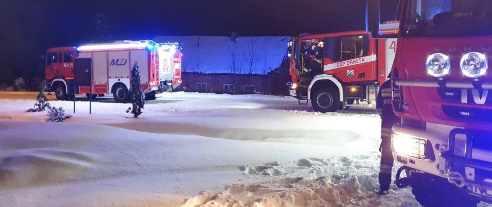 Zdjęcie przedstawia trzy samochody pożarnicze podczas interwencji. Stoją w śniegu a za nimi widoczny jest stary budynek jednorodzinny, murowany. 