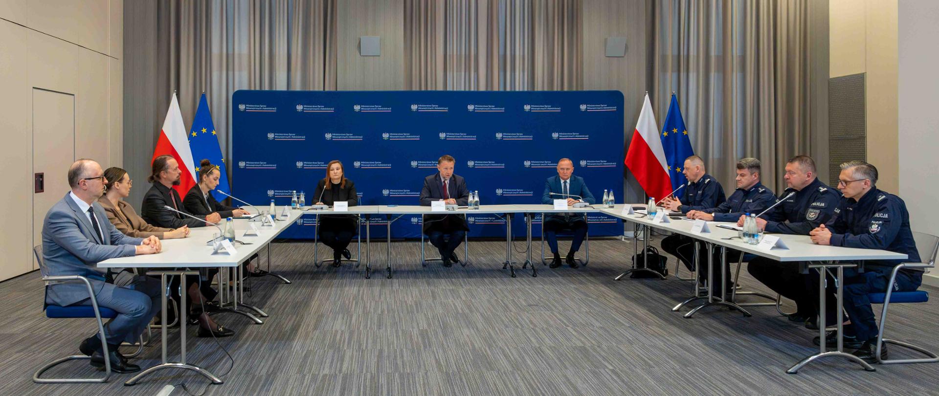 Ogólny widok sali konferencyjnej, w której uczestnicy siedzą przy dużym, rozstawionym w podkowę stole; w tle flagi Polski i Unii Europejskiej oraz ścianka z logo.