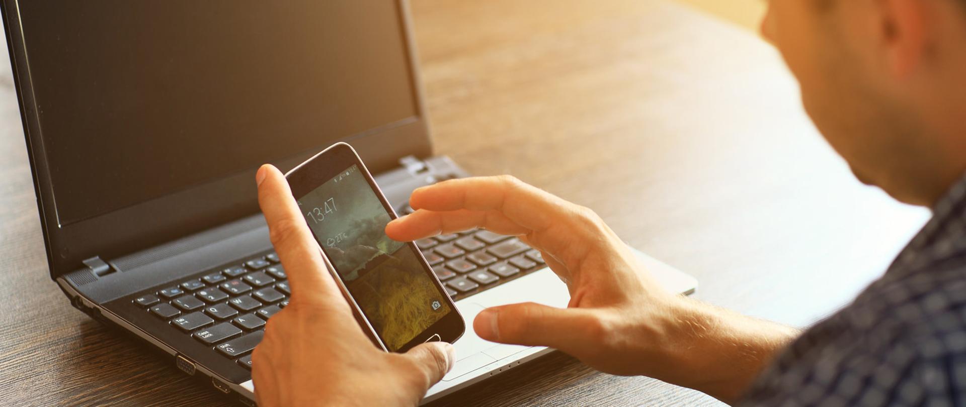 Osoba siedząca przy stole z telefonem i komputerem