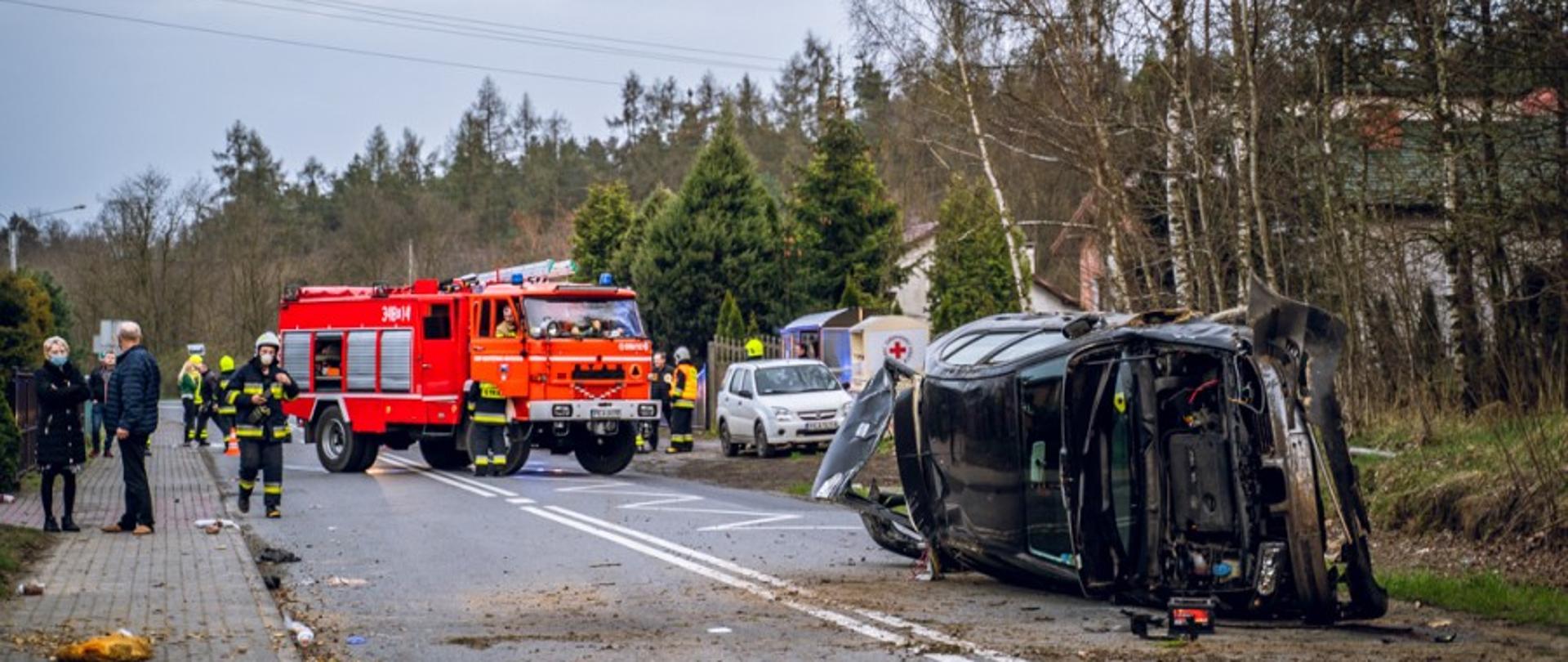 Na zdjęciu widać po prawej stronie uszkodzony samochód, leżący na jednym z boków. Wokół auta rozrzucona jest ziemia i fragmenty karoserii. Za autem widoczny las brzozowy. W centralnym miejscu widoczny jest samochód gaśniczy OSP Krzyżówki. Auto w kolorze czerwonym, z oznaczeniami na drzwiach i sygnalizacją świetlną na dachu. Po lewej stronie zdjęcia widoczne są postaci ludzi i ratowników stojących na chodniku i jezdni. Za samochodem straży pożarnej widać zielony las iglasty. Na prawo od auta jest wiata przystankowa, kontener na odzież używaną i białe auto. 