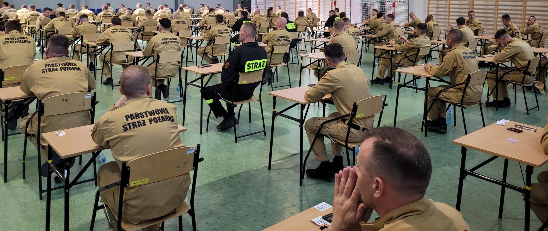 Sala gimnastyczna SA PSP w Krakowie. Widok z góry. Wewnątrz w ustawionych w rzędy ławkach siedzą zdający egzamin strażacy. 
