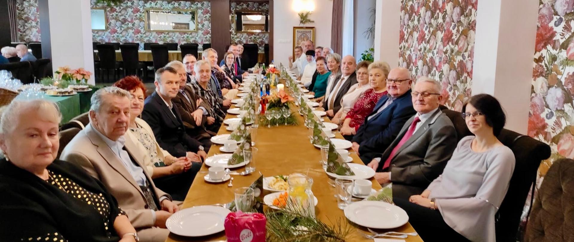 Zdjęcie przedstawia zaproszonych gości przy długim zastawionym stole. W tle widać salę balowa.