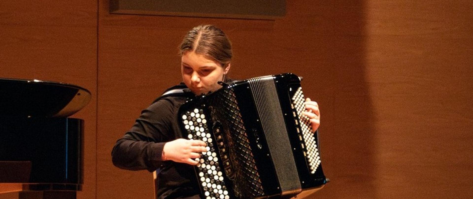 Na zdjęciu widoczna jest młoda dziewczyna siedząca na krześle i grająca na akordeonie. Ma ciemne włosy związane z tyłu głowy i ubrana jest w ciemne ubrania. Obok niej znajduje się fragment czarnego fortepianu. Scena ma drewnianą podłogę i tło.