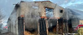 Widok budynku z czoła z otwartymi wrotami oraz wydobywającym się dymem z pomieszczenia przez jeden z otworów widać spaloną więźbę dachową, przed budynkiem materiały osunięte z pomieszczenia