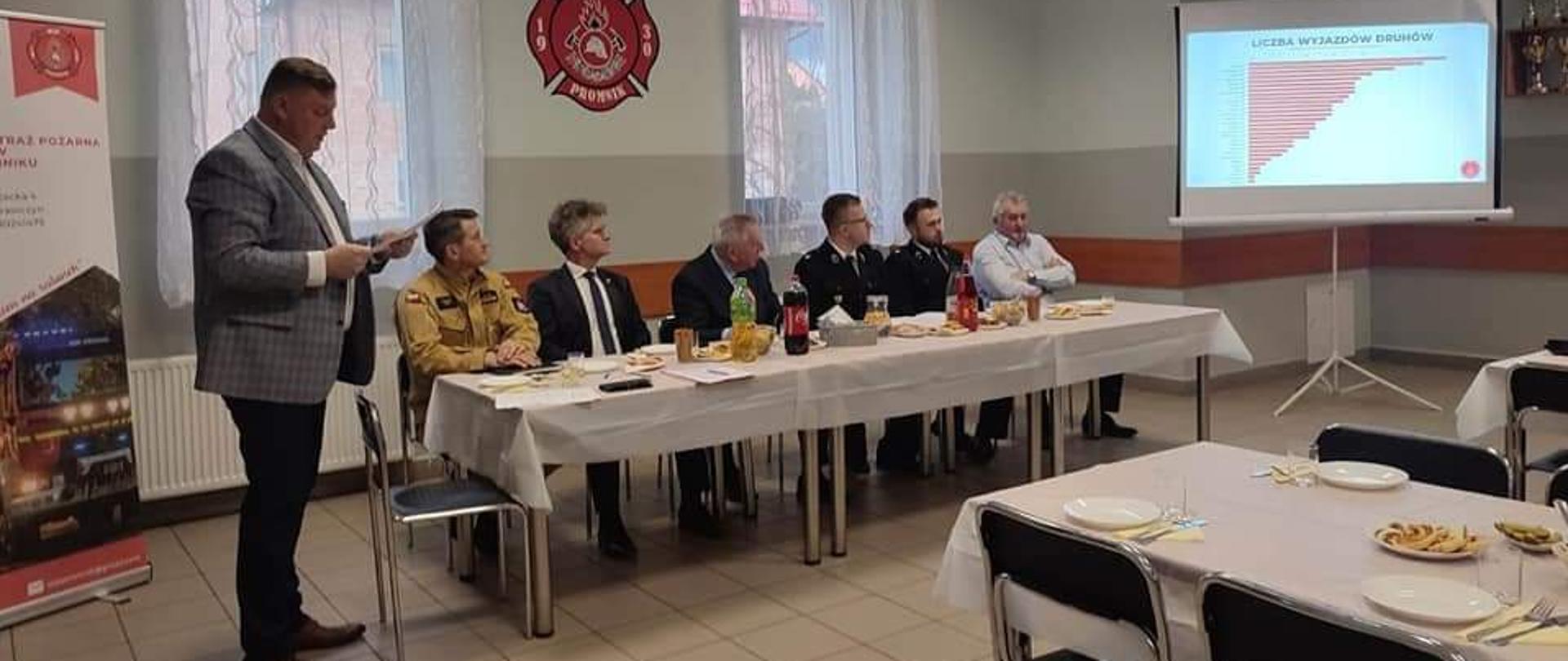 Zdjęcie przedstawia gości i organizatorów spotkania sprawozdawczego Ochotniczej Straży Pożarnej w Promniku podczas prezentacji.