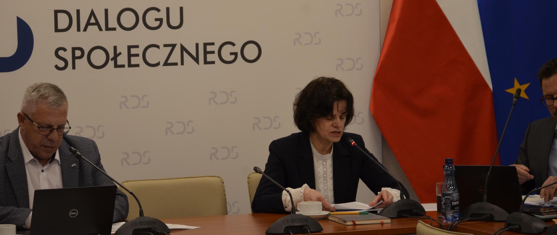 Na zdjęciu widnieje stół prezydialny za którym siedzi Przewodniczący Zespołu ds. budżetu, wynagrodzeń i świadczeń socjalnych – Leszek Miętek a po Jego lewej Hanna Majszczyk – Podsekretarz Stanu w Ministerstwie Finansów. Po lewej stronie widnieje flaga UE. W tle siedzących znajduje się baner z napisem Rada Dialogu Społecznego.
