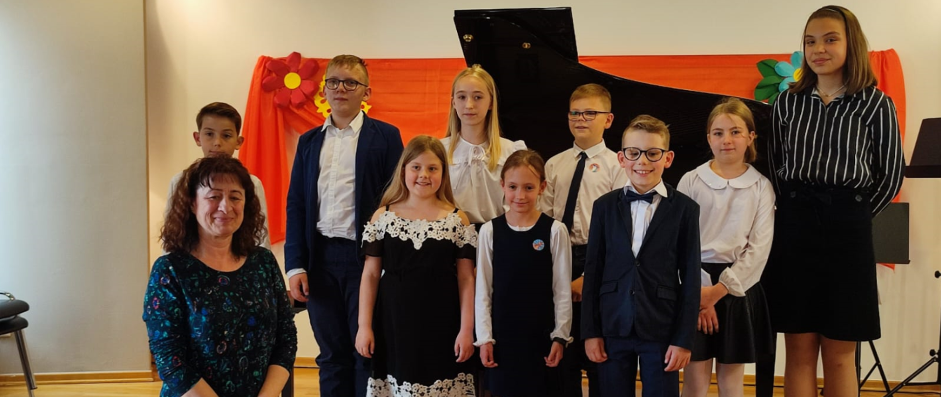Na zdjęciu grupa uczniów wraz z nauczycielką- brunetka w granatowej sukience w kwiatki. W tle otwarty fortepian. Wszyscy stoją na scenie. Uczniowie: dziewczyny i chłopcy ubrani odświętnie.